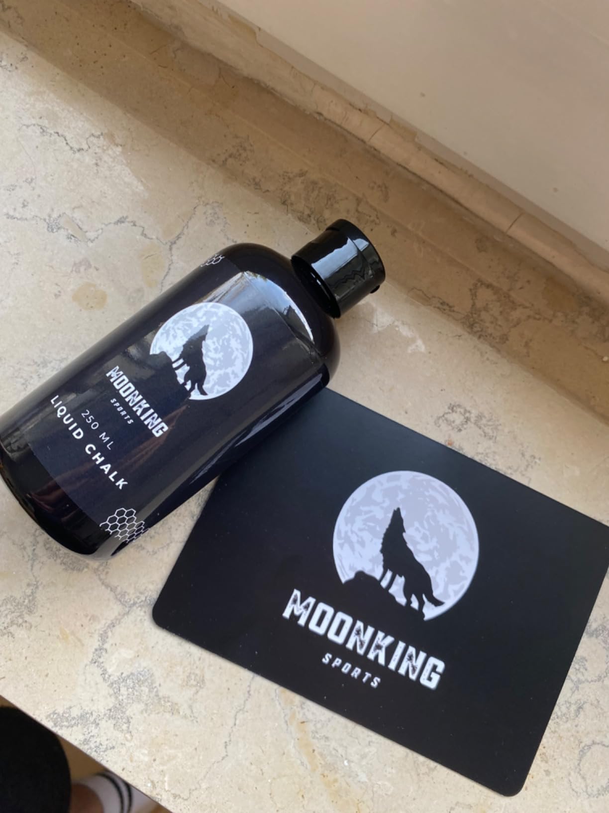 Eine Flasche Flüssigkreide mit der Aufschrift „MOONKING SPORTS“ liegt auf einer Marmoroberfläche neben einer Karte mit der Silhouette eines Wolfs vor einem Vollmond, ebenfalls mit dem Logo von MOONKING SPORTS.