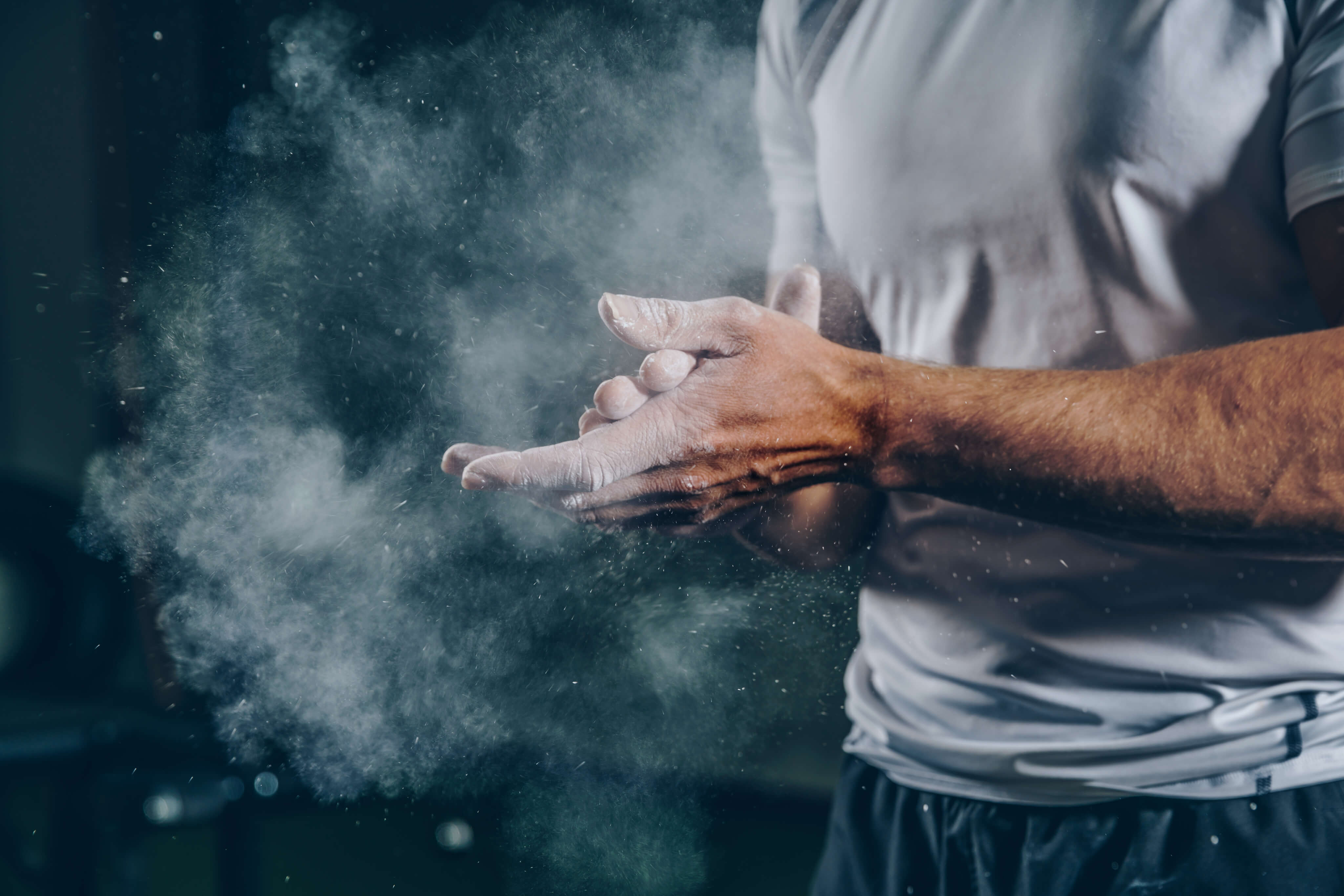 Eine Person in weißem T-Shirt und dunklen Shorts klatscht in die Hände und erzeugt eine Wolke aus weißem Kreidestaub vor einem dunklen Hintergrund. Das Bild vermittelt ein Gefühl von Vorbereitung und Konzentration, wie man es häufig im Sport oder im Fitnessstudio sieht.